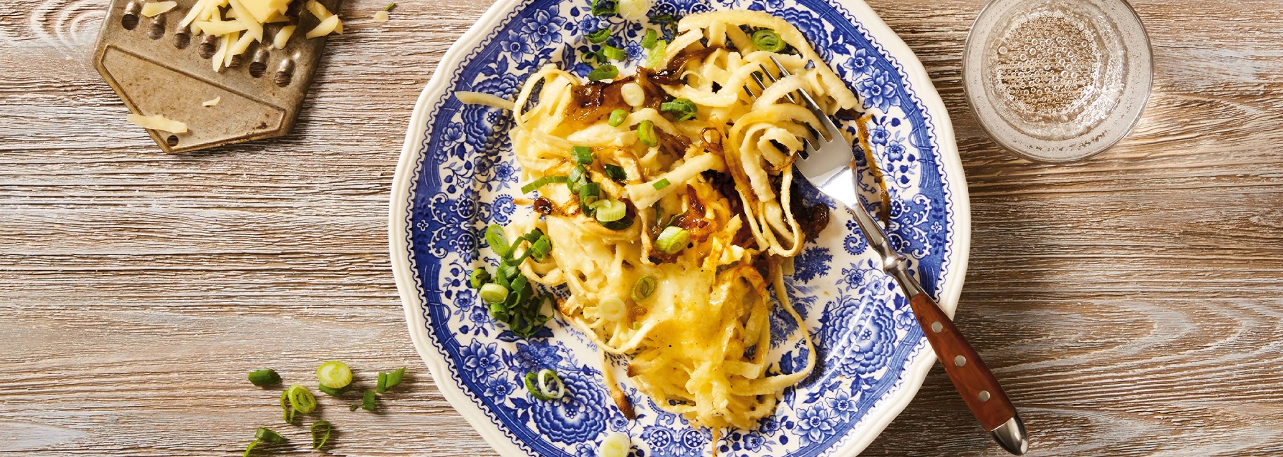 En tallerken med ostet spätzle toppet med karamelliserede løg og forårsløg, ved siden af en rivejern med revet ost og et glas vand.