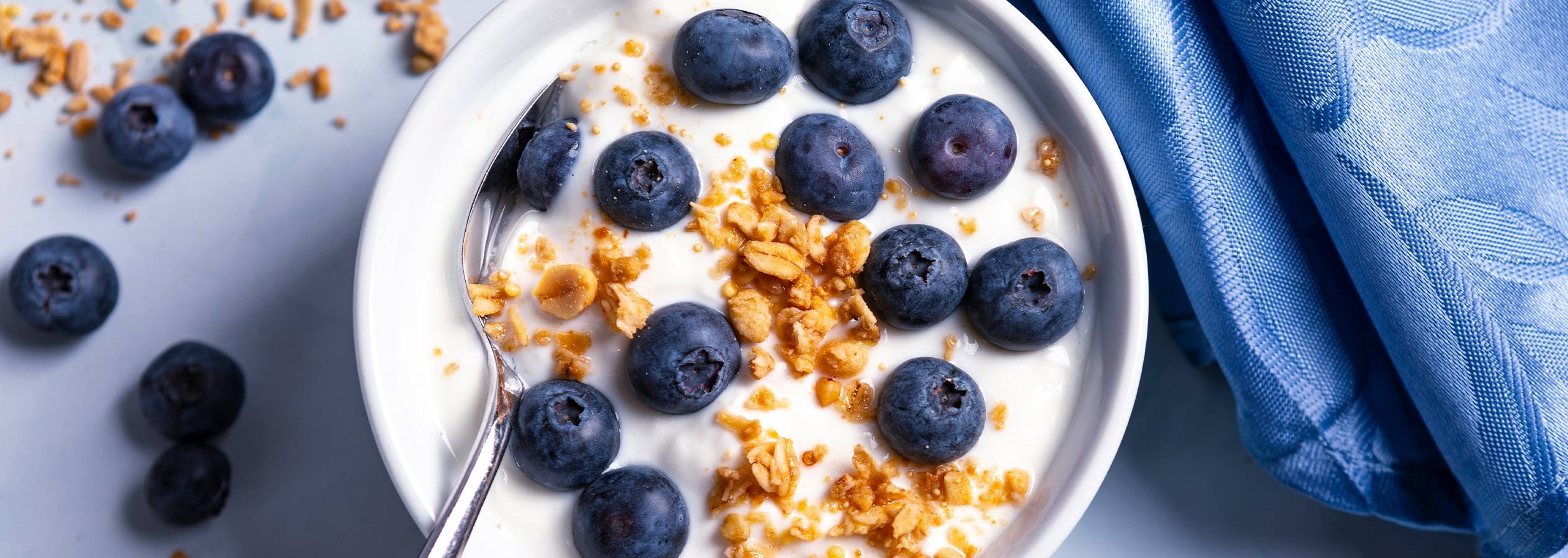 En skål med yoghurt, blåbær og granola med en ske og et blåt klæde.