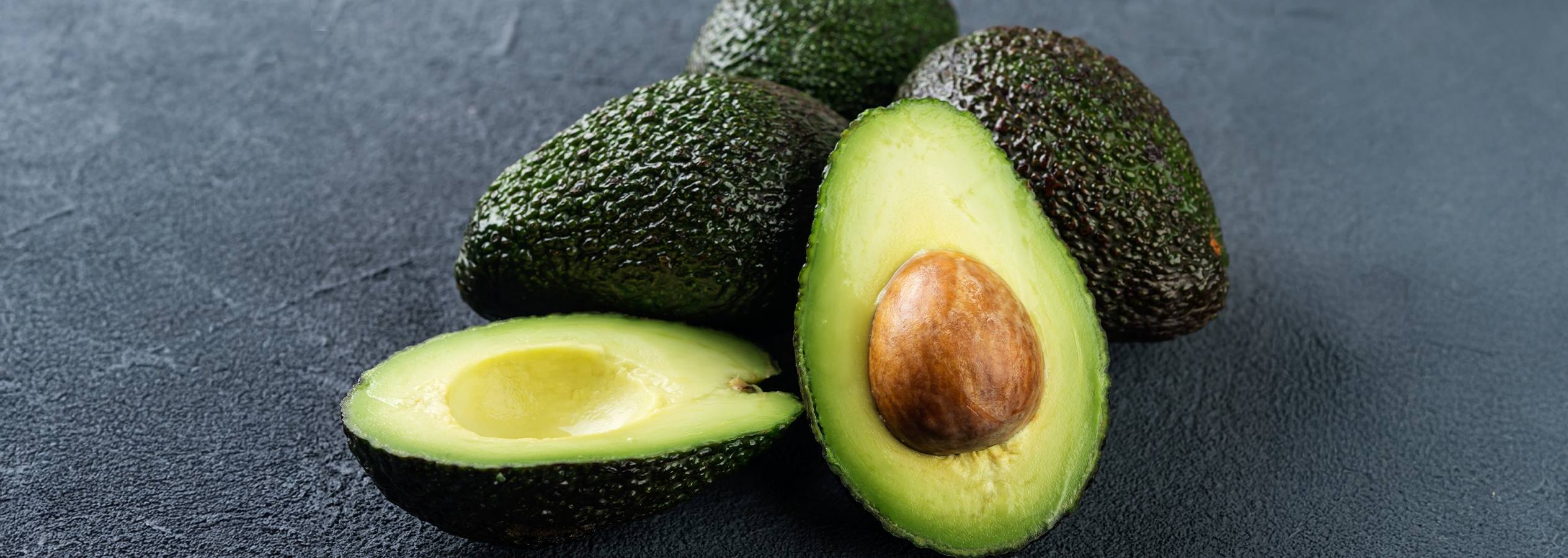 Hele og halverede avocadoer med en kerne på en mørk overflade.