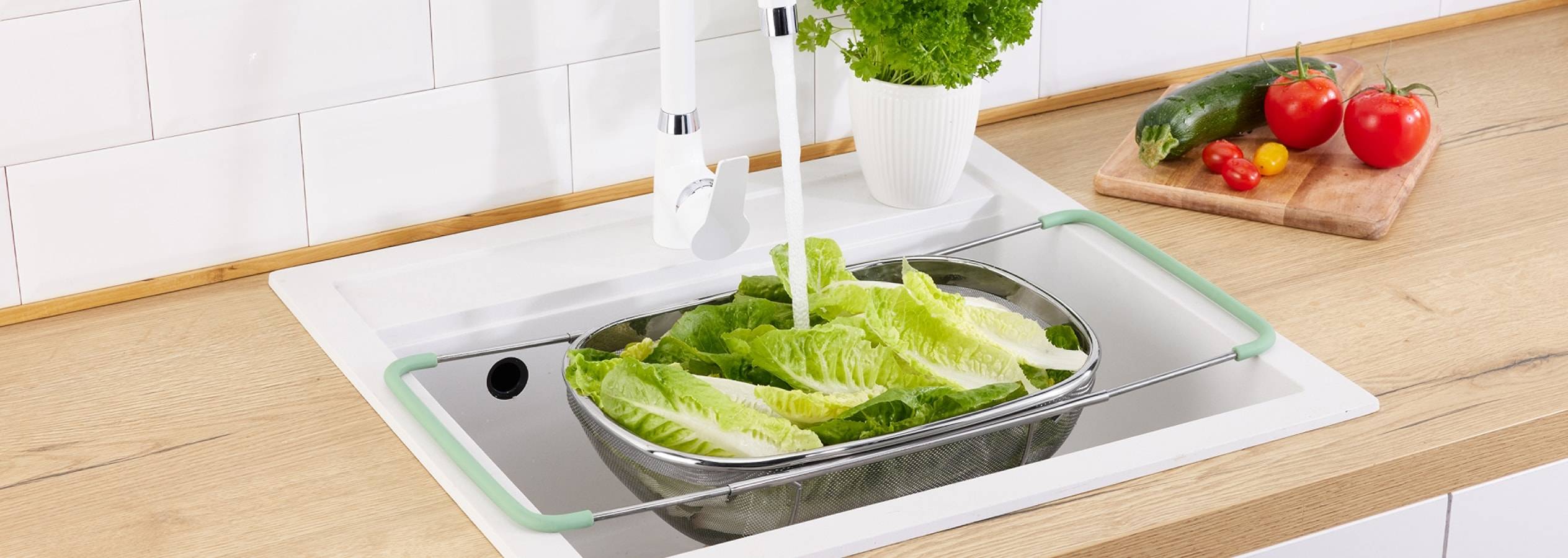 Et køkken med et afløbsstativ i rustfrit stål med salat i, der vaskes under en vandhane.