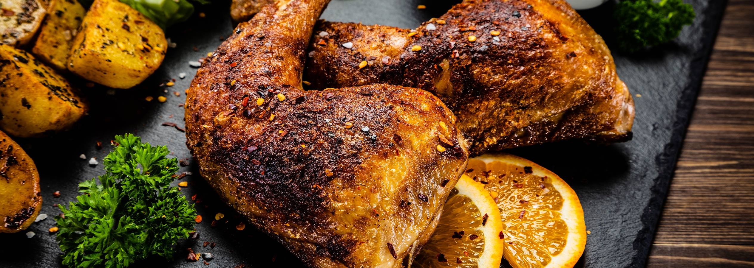 Grillet kylling med kartofler, persille og appelsinskiver på en sort skiferplade.