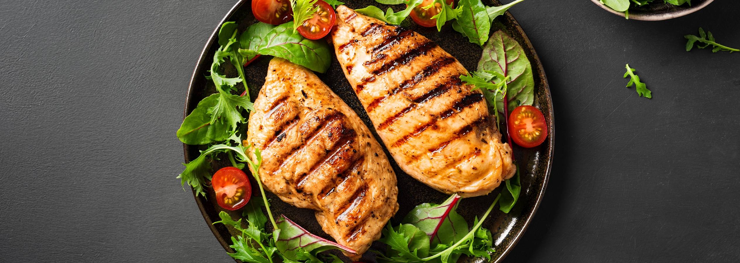Grillet kyllingebryst med salat og cherrytomater på en mørk tallerken.