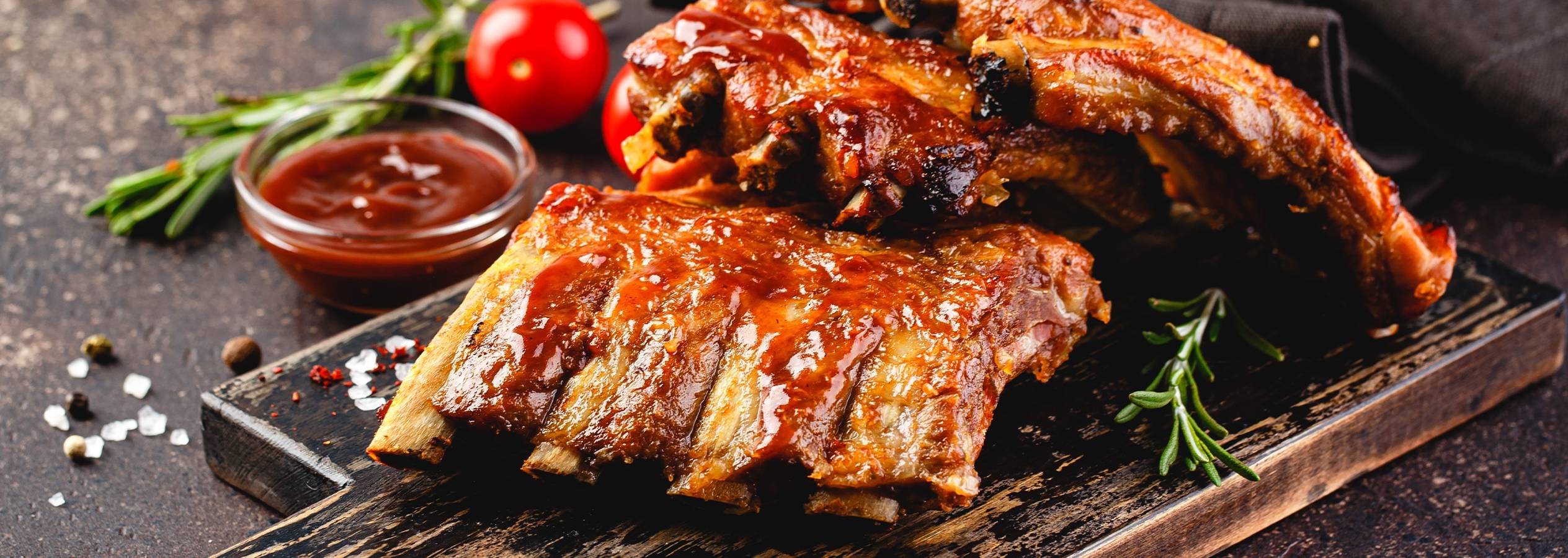 Grillede spareribs med barbecuesauce, rosmarin og tomater på et træbræt.