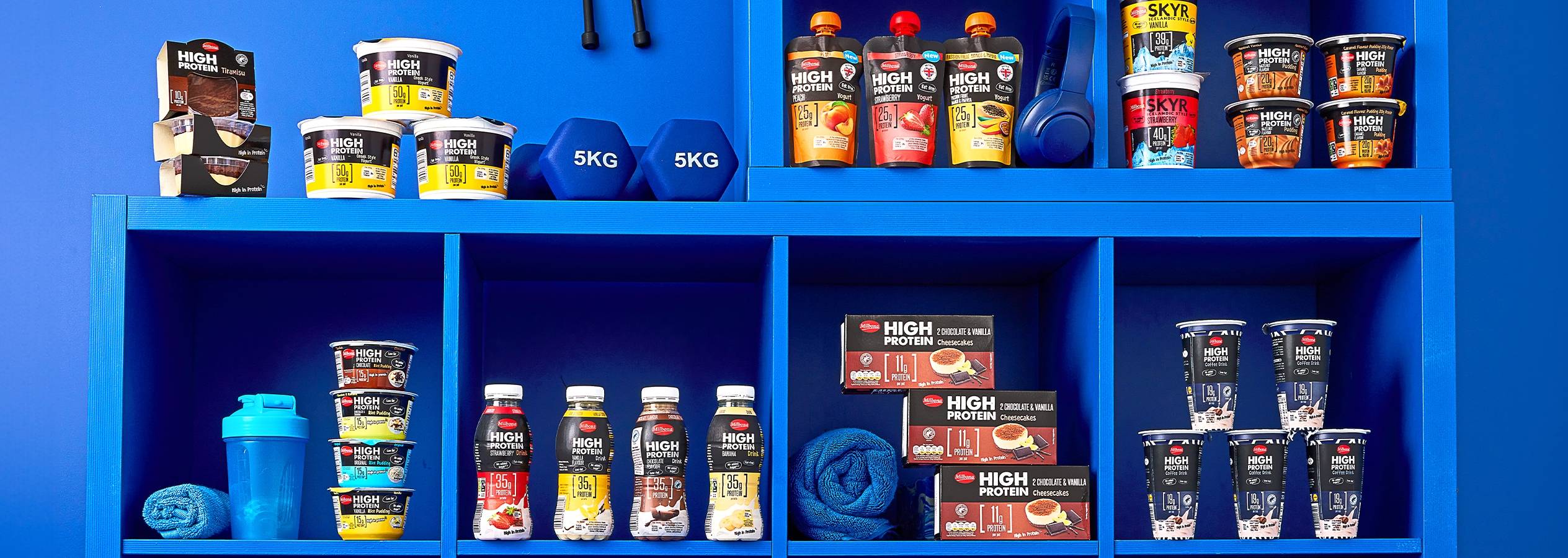 En hylde fyldt med forskellige proteinrige produkter, herunder yoghurt, buddinger, drikkevarer og desserter.