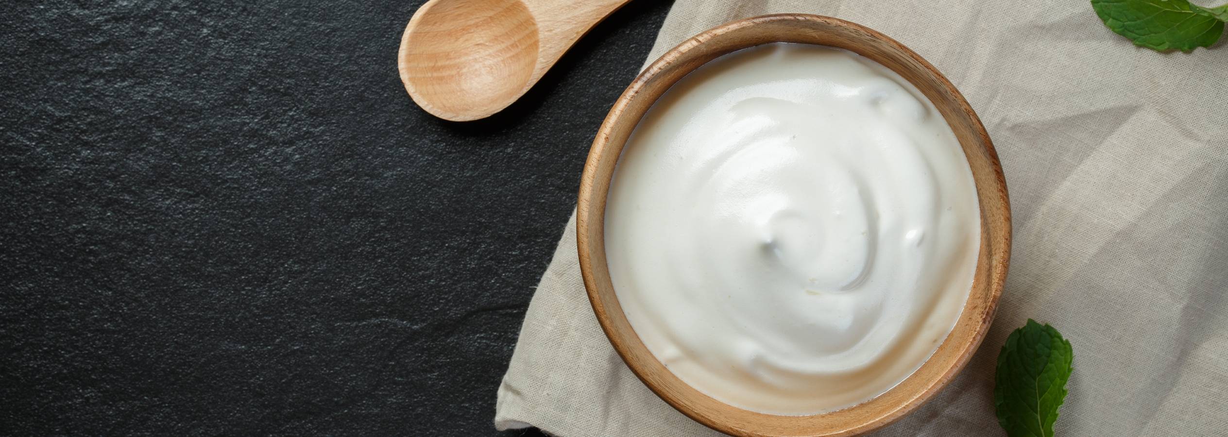 En skål med yoghurt, en træske og mynteblade på en mørk overflade.