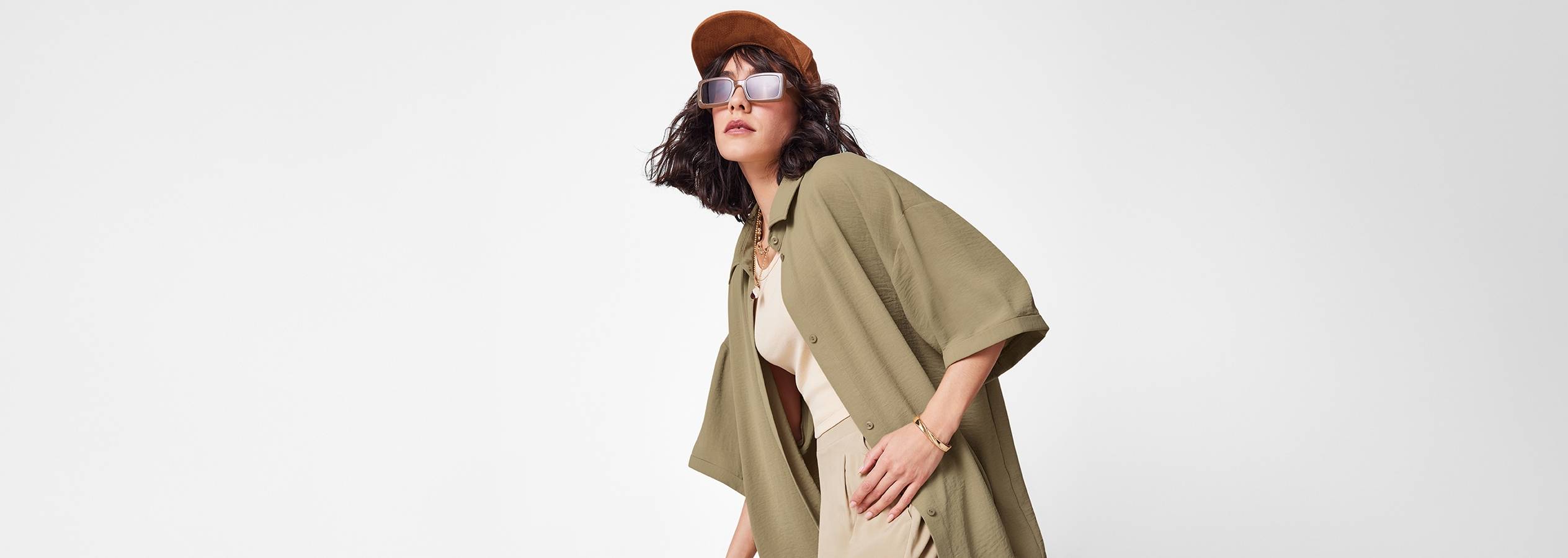 Model iført en olivengrøn skjorte, beige bukser, solbriller og en brun kasket.