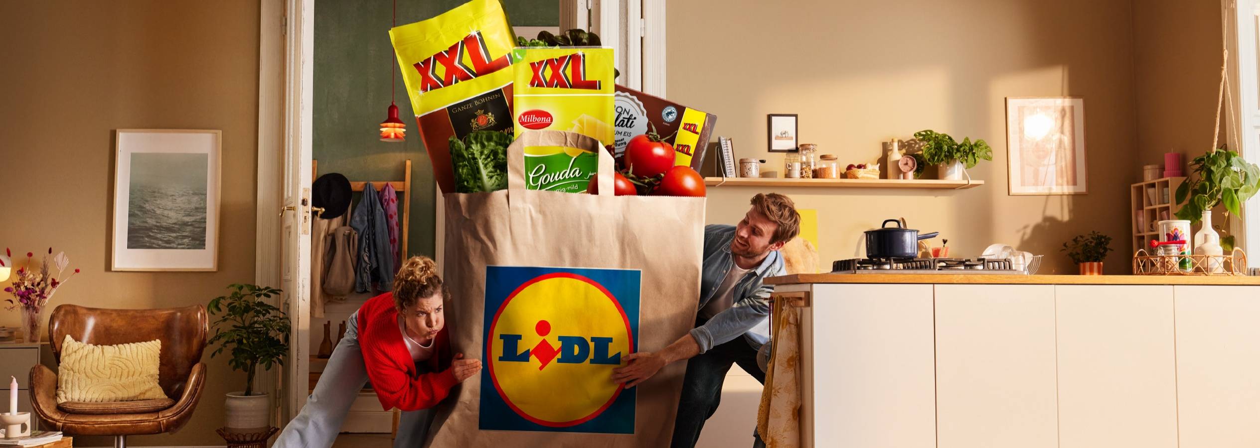 To personer bærer en overdimensioneret Lidl-indkøbspose fyldt med dagligvarer som grøntsager, ost og kaffe.
