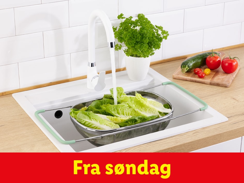 Et grøntsagsbakke i rustfrit stål med lysegrønne håndtag vaskes i en køkkenvask.