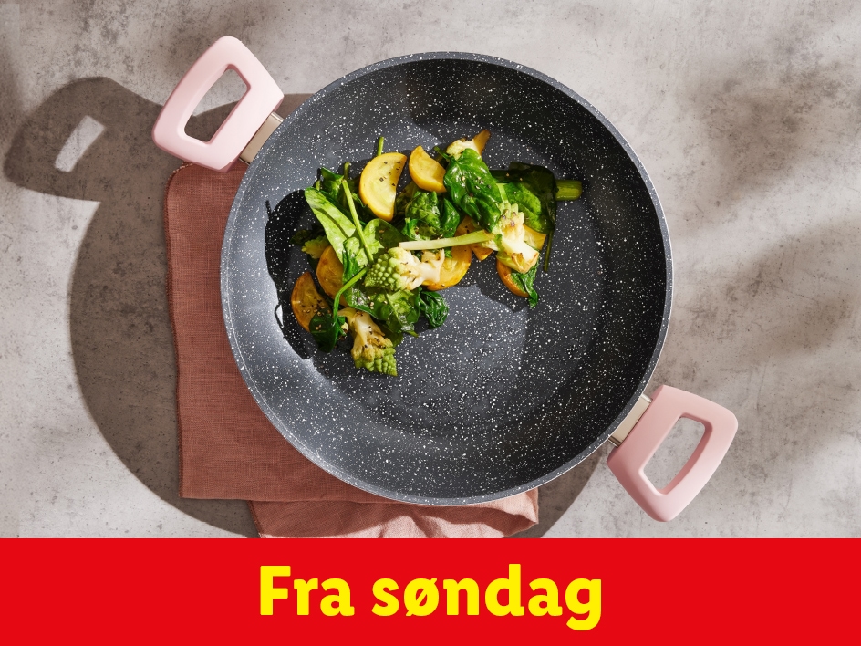 En sort stegepande med lyserøde håndtag fyldt med grøntsager, på et gråt underlag.