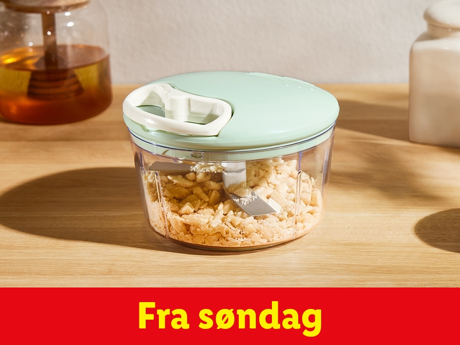 En manuel hakker fyldt med hakkede nødder på et træbord, med en honningkrukke i baggrunden.