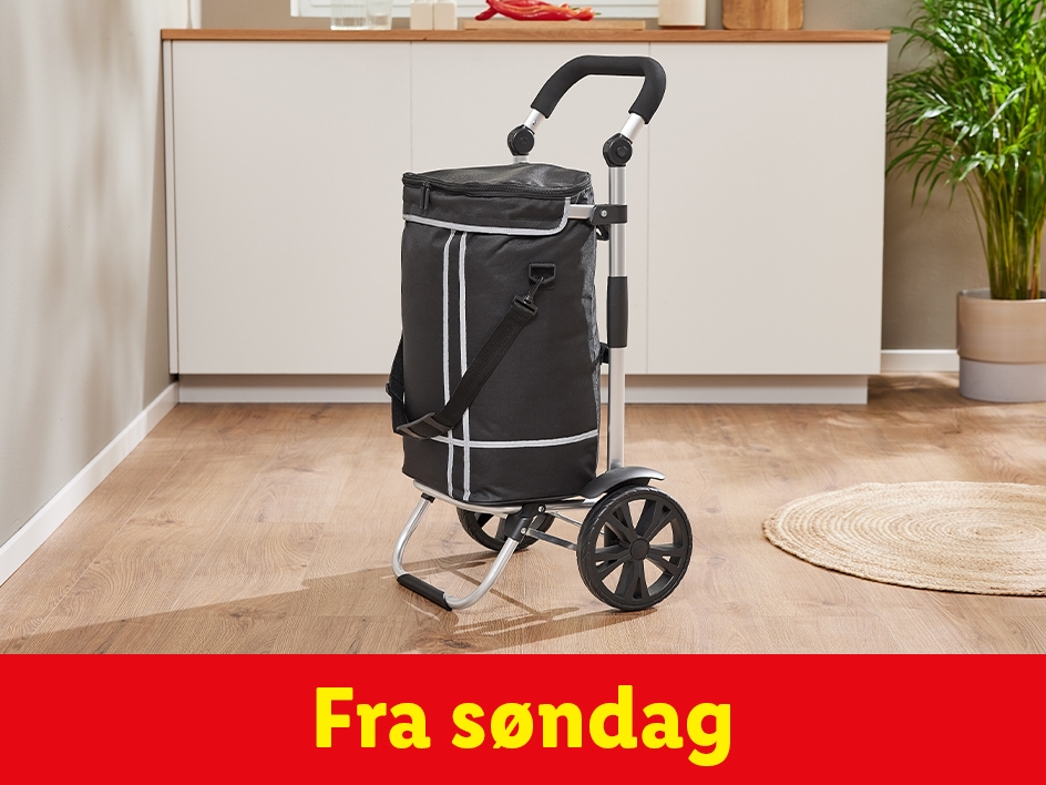 En sort indkøbsvogn med hjul står på et trægulv i et køkken.