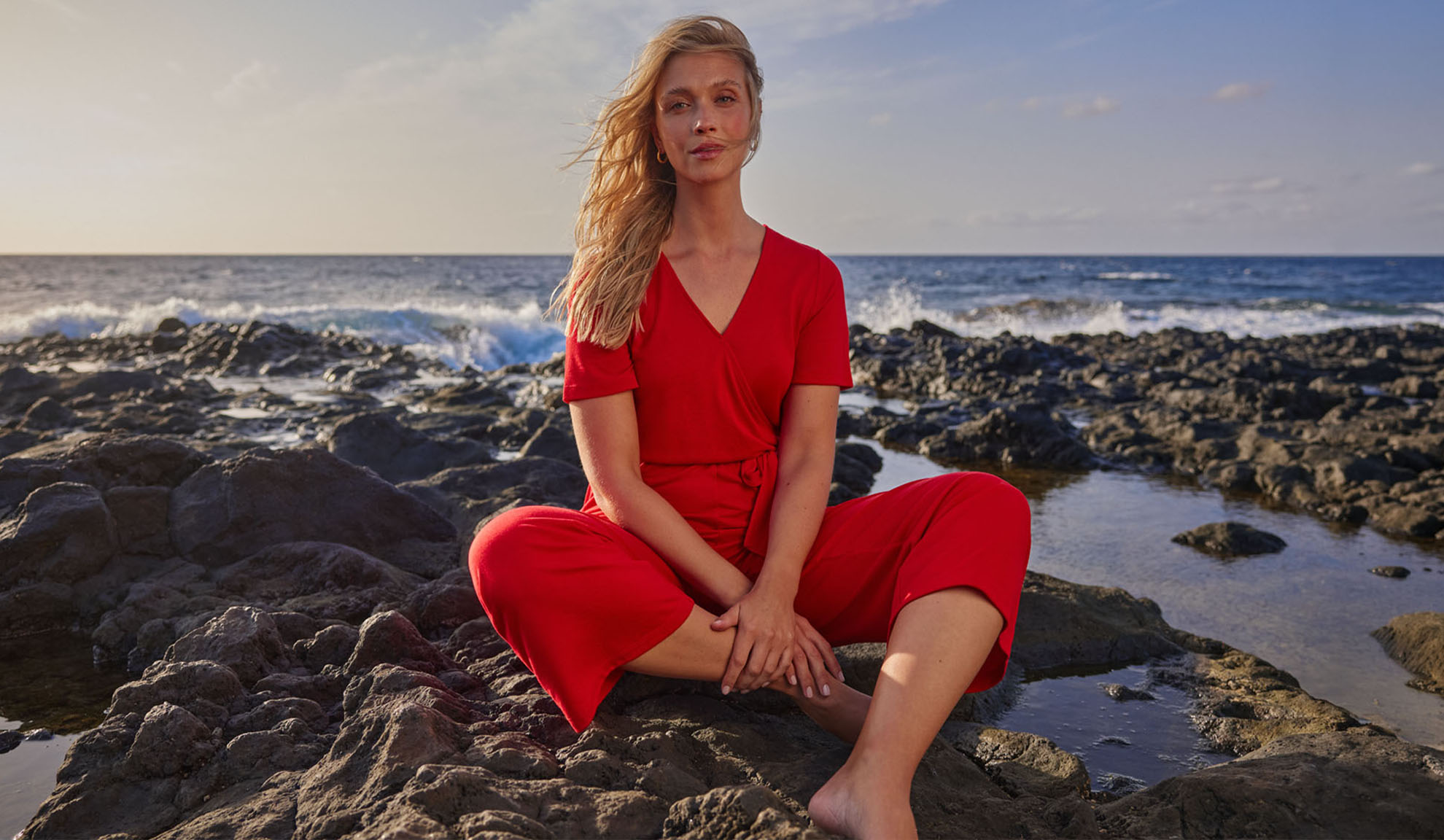 Kvinde i rød jumpsuit sidder på klipper ved havet.