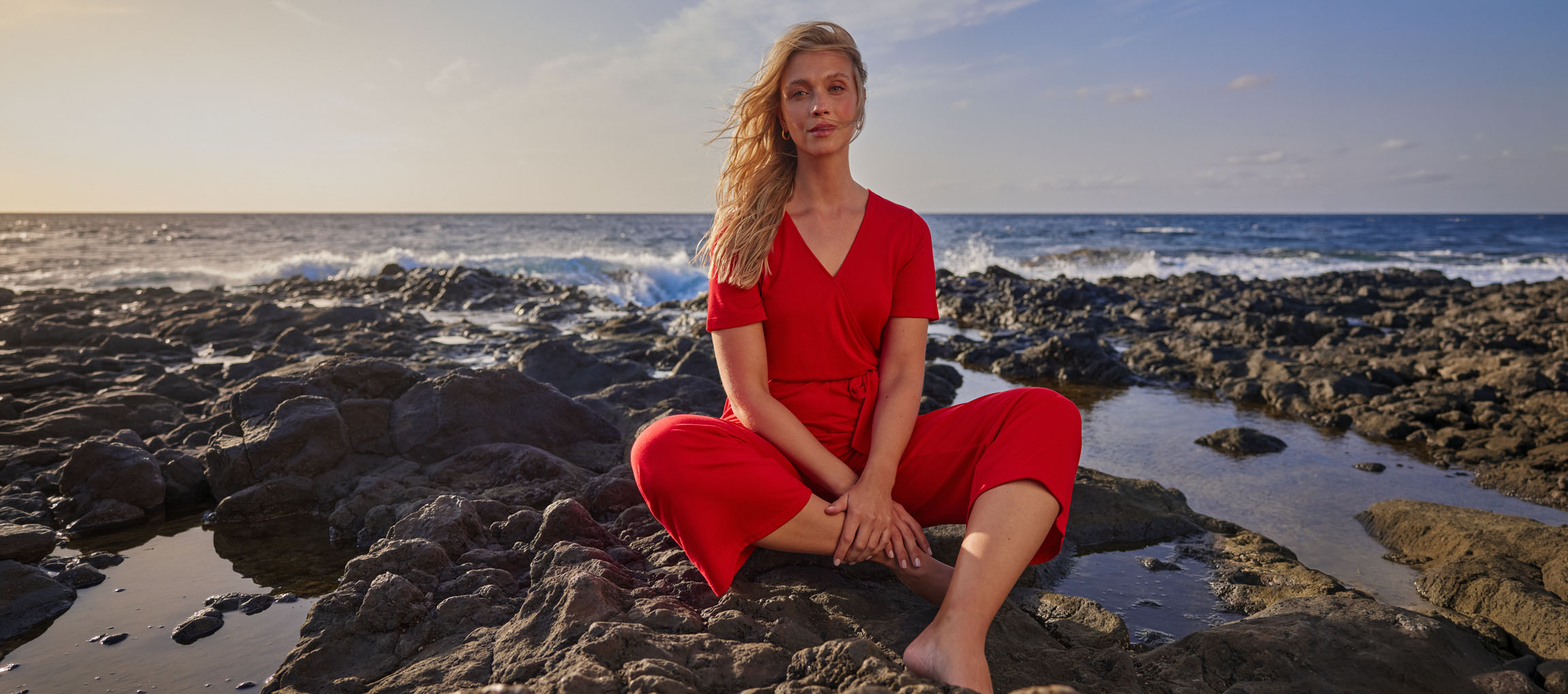 Kvinde i rød jumpsuit sidder på en stenstrand ved havet.