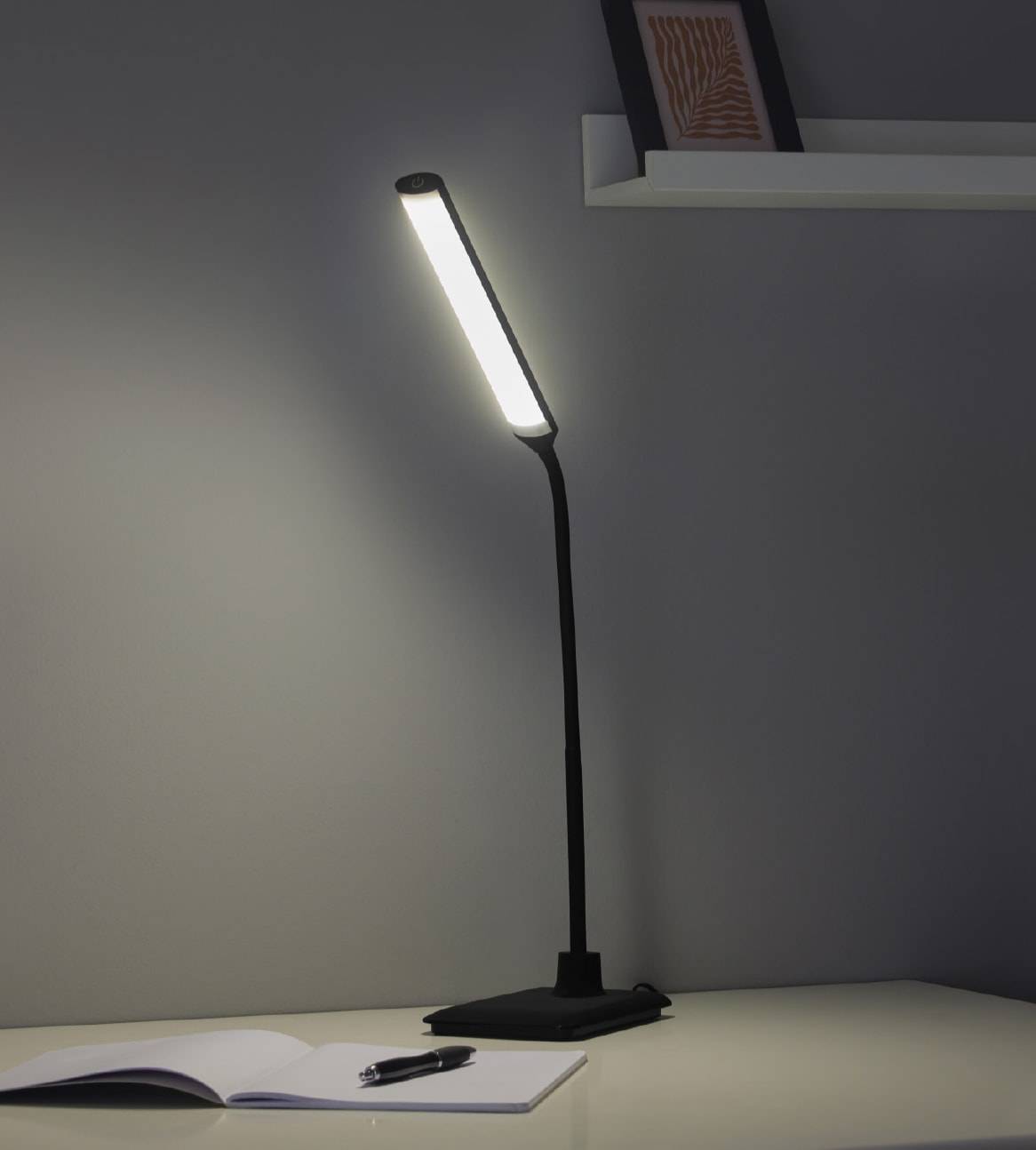 En sort LED skrivebordslampe med et langt, tyndt lyspanel lyser op på et skrivebord med en notesbog og en pen.
