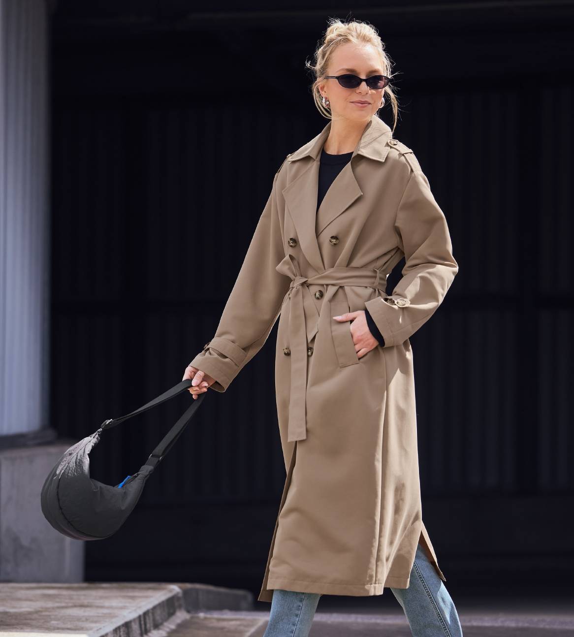 En kvinde i en beige trenchcoat, jeans og solbriller holder en sort taske.