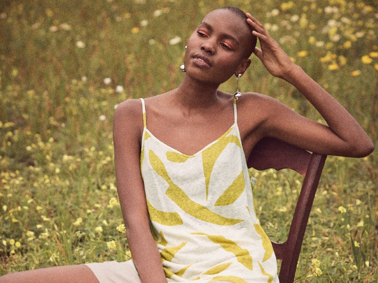 En model iført en sommerlig, gul-og-hvid tanktop sidder på en stol i et blomsterfyldt felt.