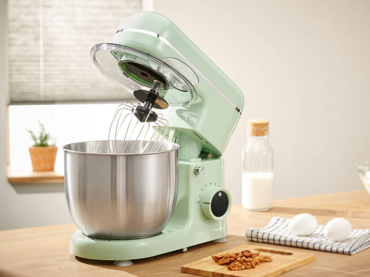 Mintgrøn standmixer med piskeris og skål, klar til bagning med mælk, æg og nødder på et træbord.