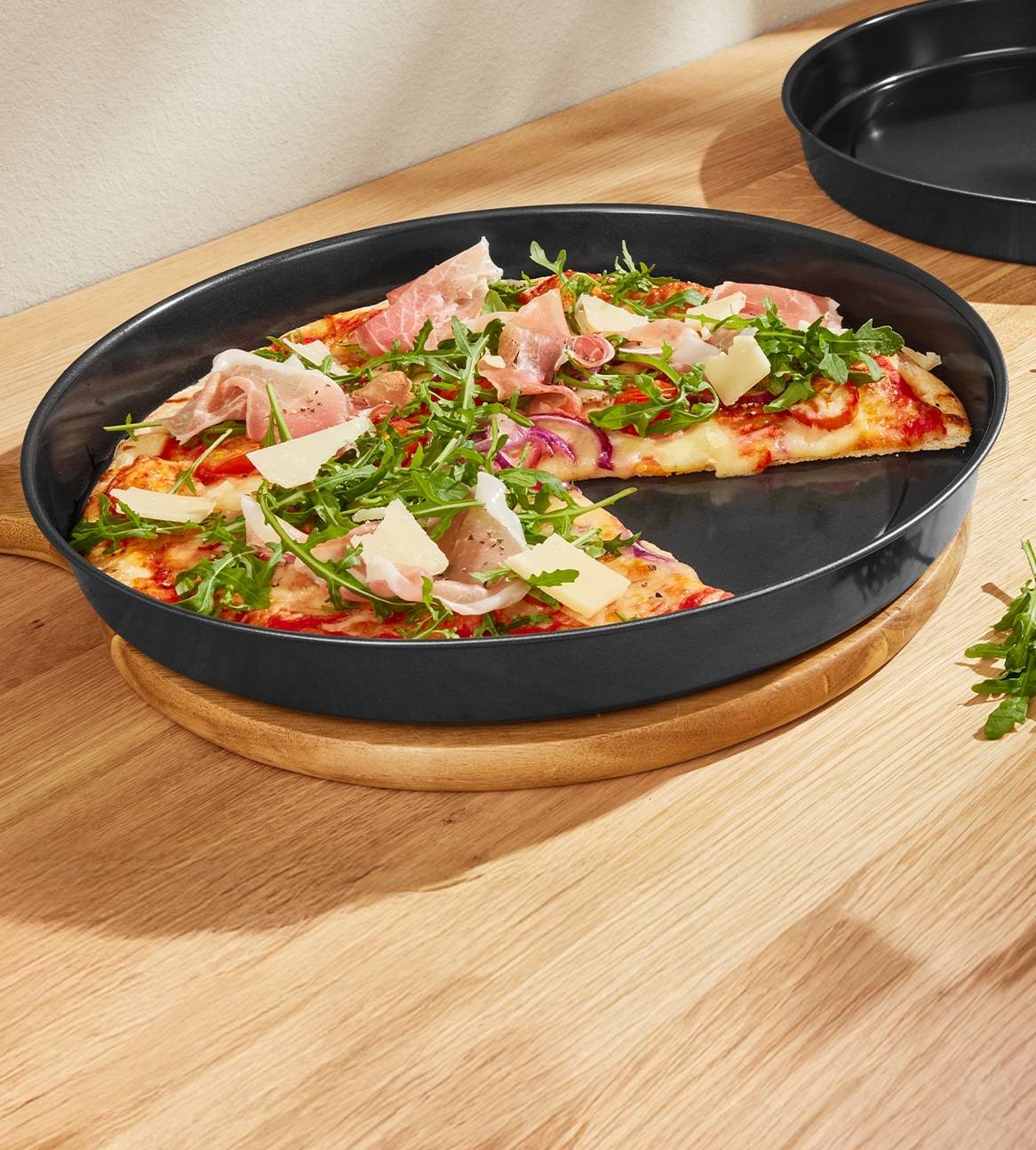 Pizza med parmaskinke, rucola og parmesan i en sort bageform på et træbord.