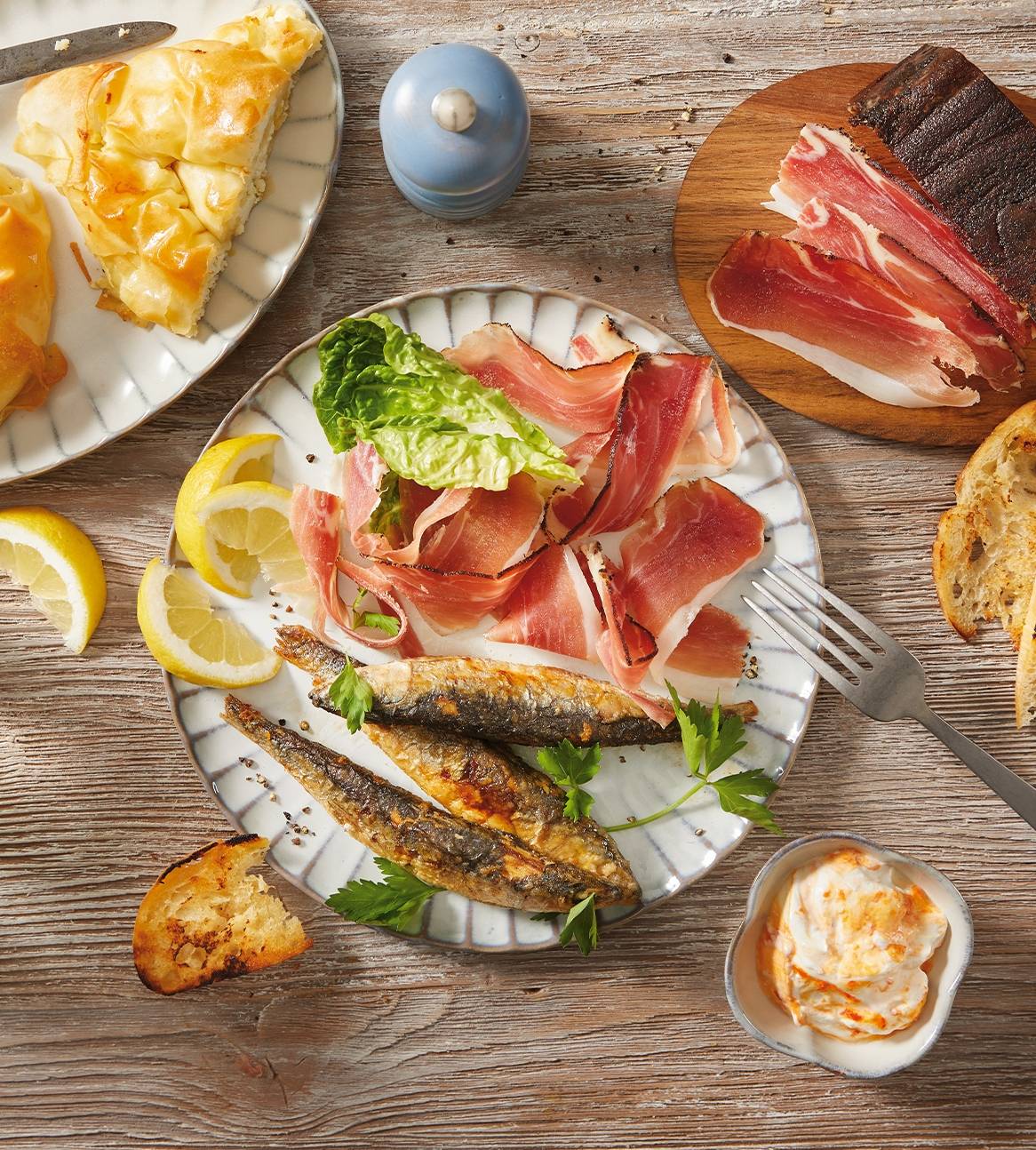 En tallerken med stegte fisk, skinke og citroner, med en tærte og brød på et træbord.