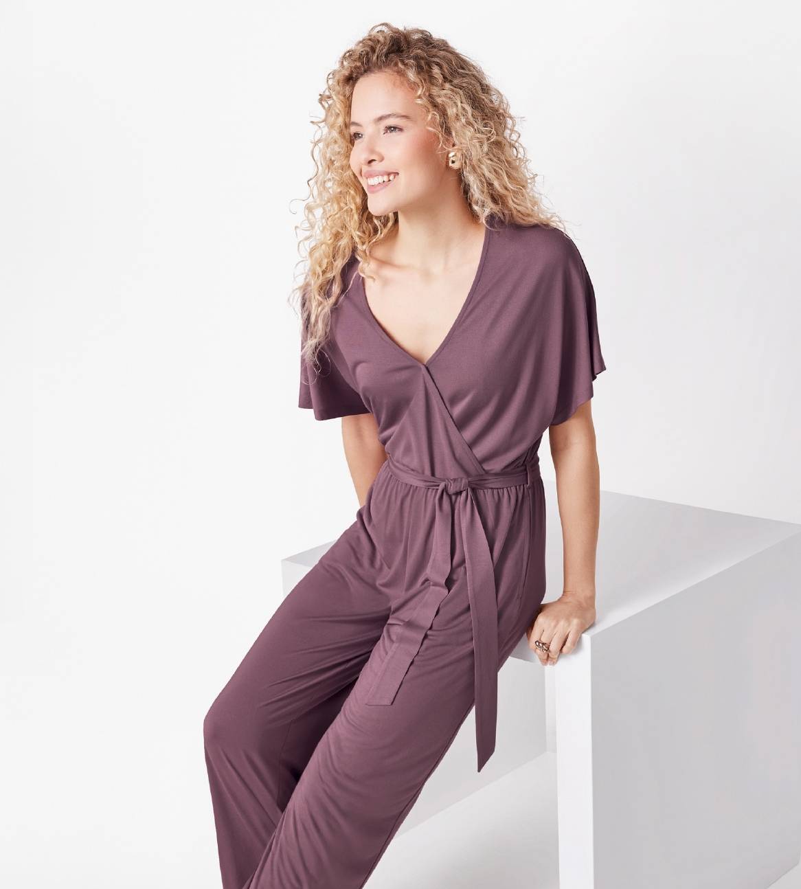 En model viser en mørklilla jumpsuit med V-hals og bælte.