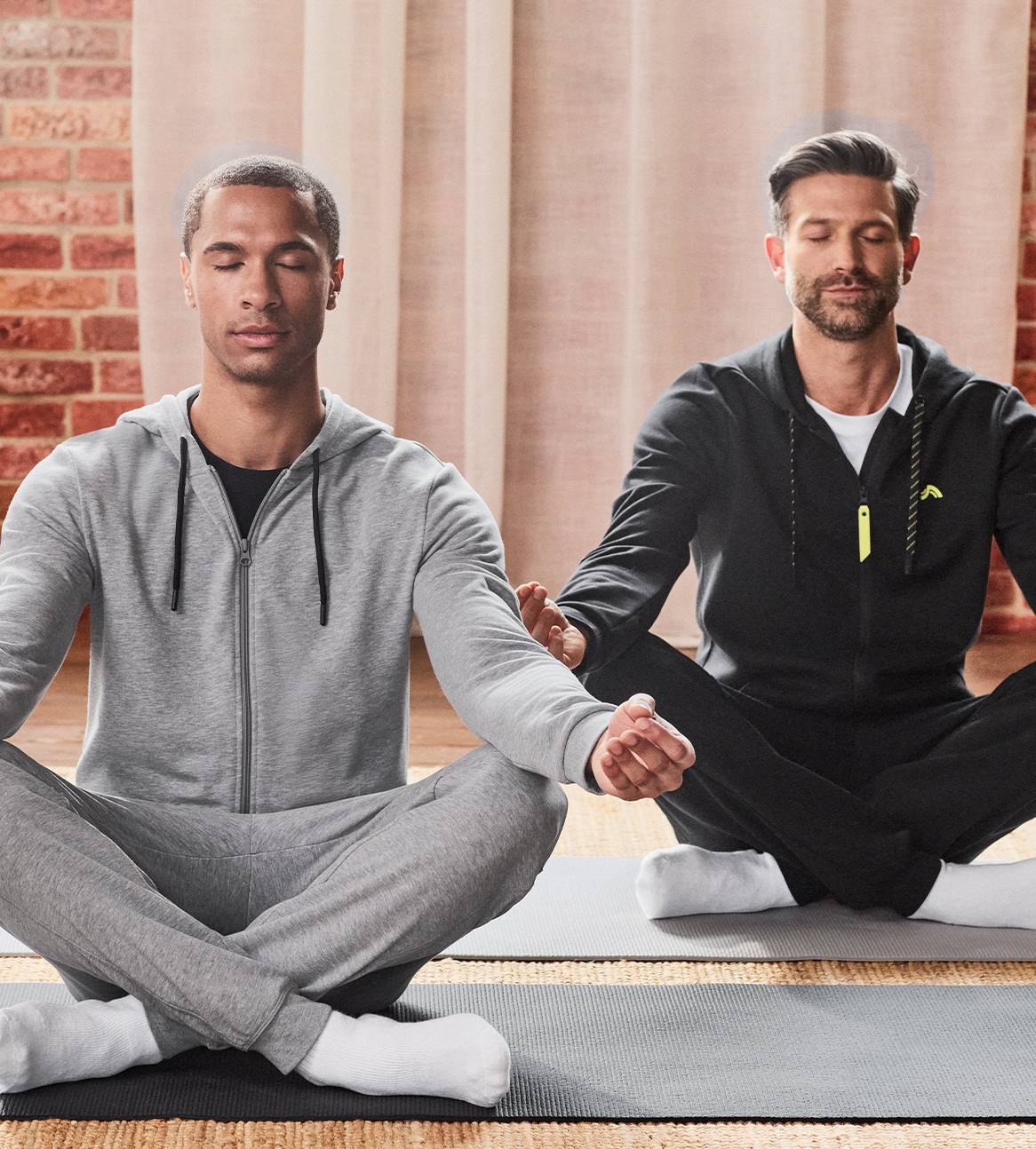 To mænd i grå og sorte træningsdragter med hætter mediterer på yogamåtter.