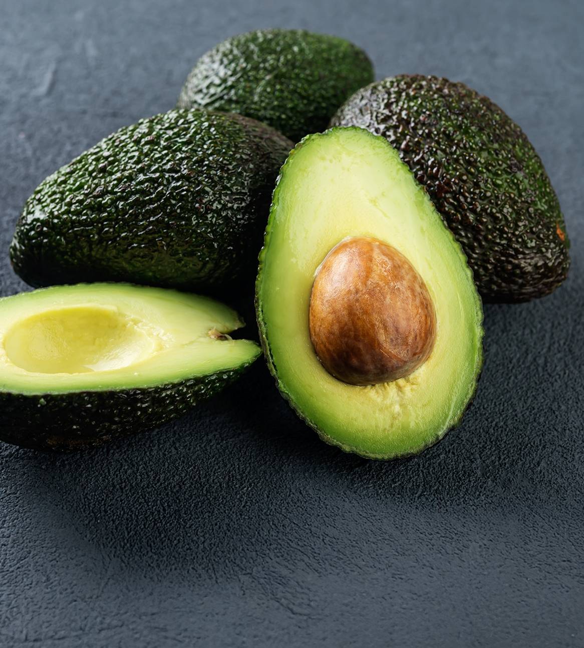 Hele og halverede avocadoer med sten på en mørk overflade.