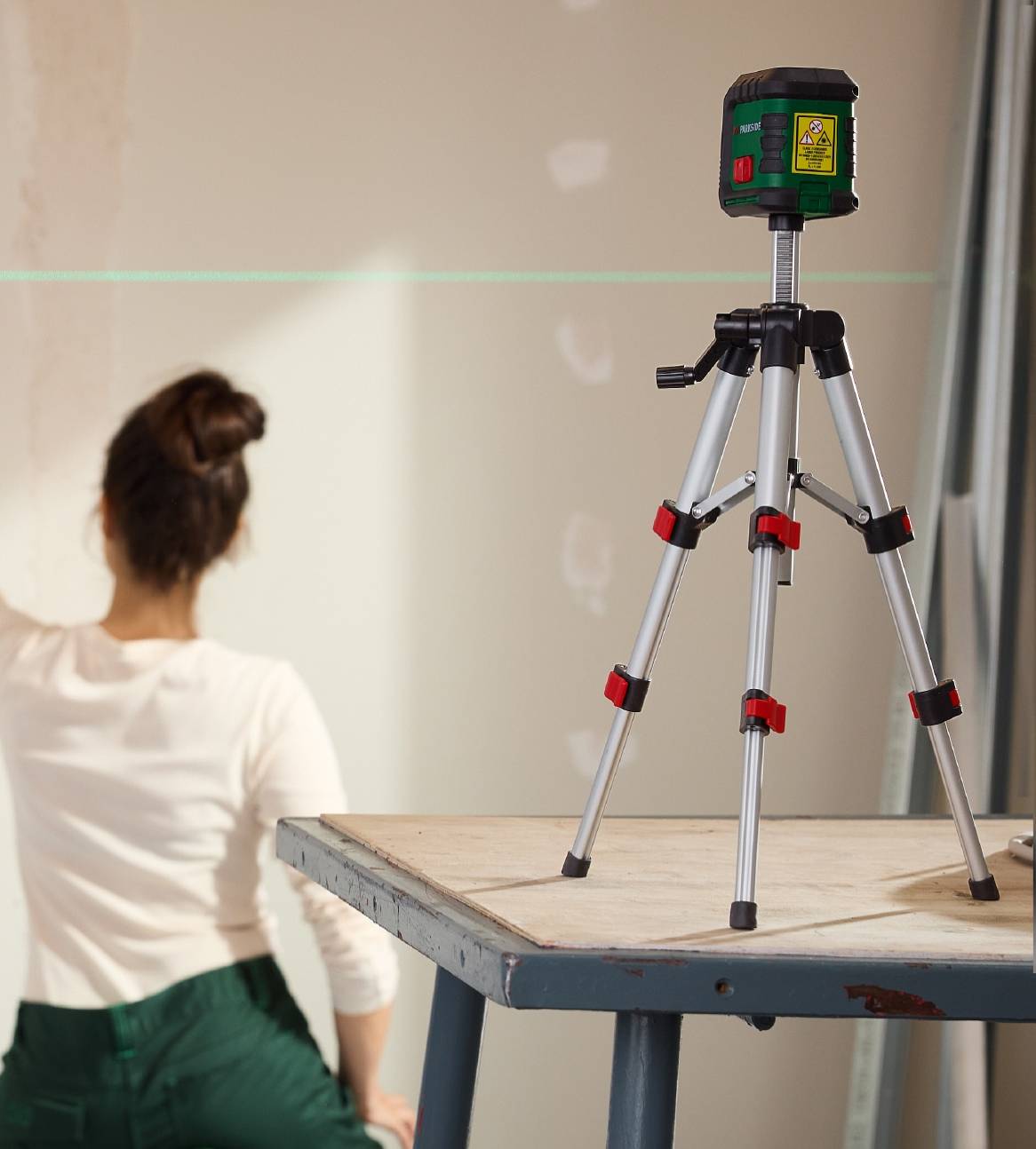 En grøn lasermåler på et stativ projicerer en grøn linje på en væg, hvor en person arbejder.