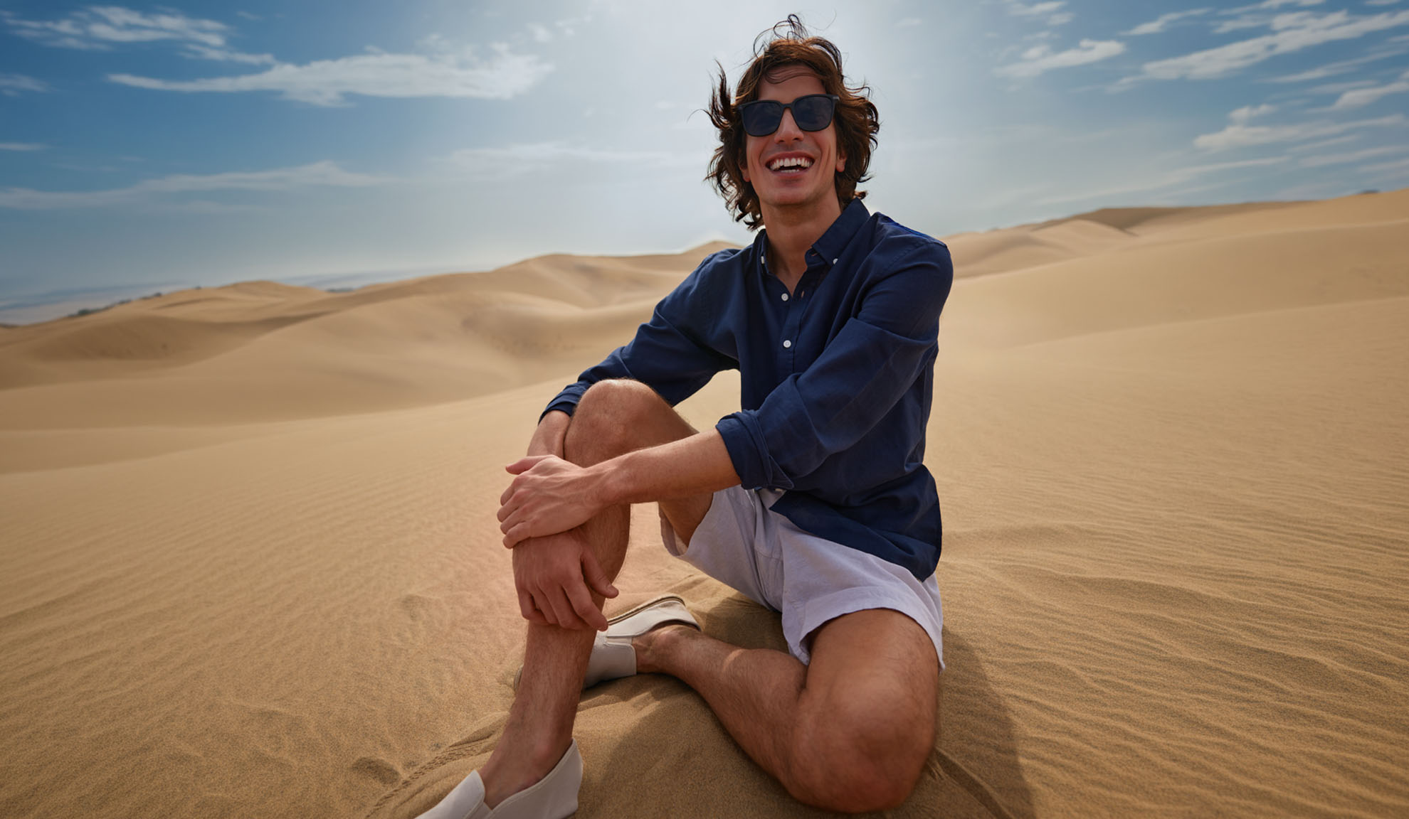 Mand i mørkeblå skjorte og lyse shorts sidder smilende på sandklitter.