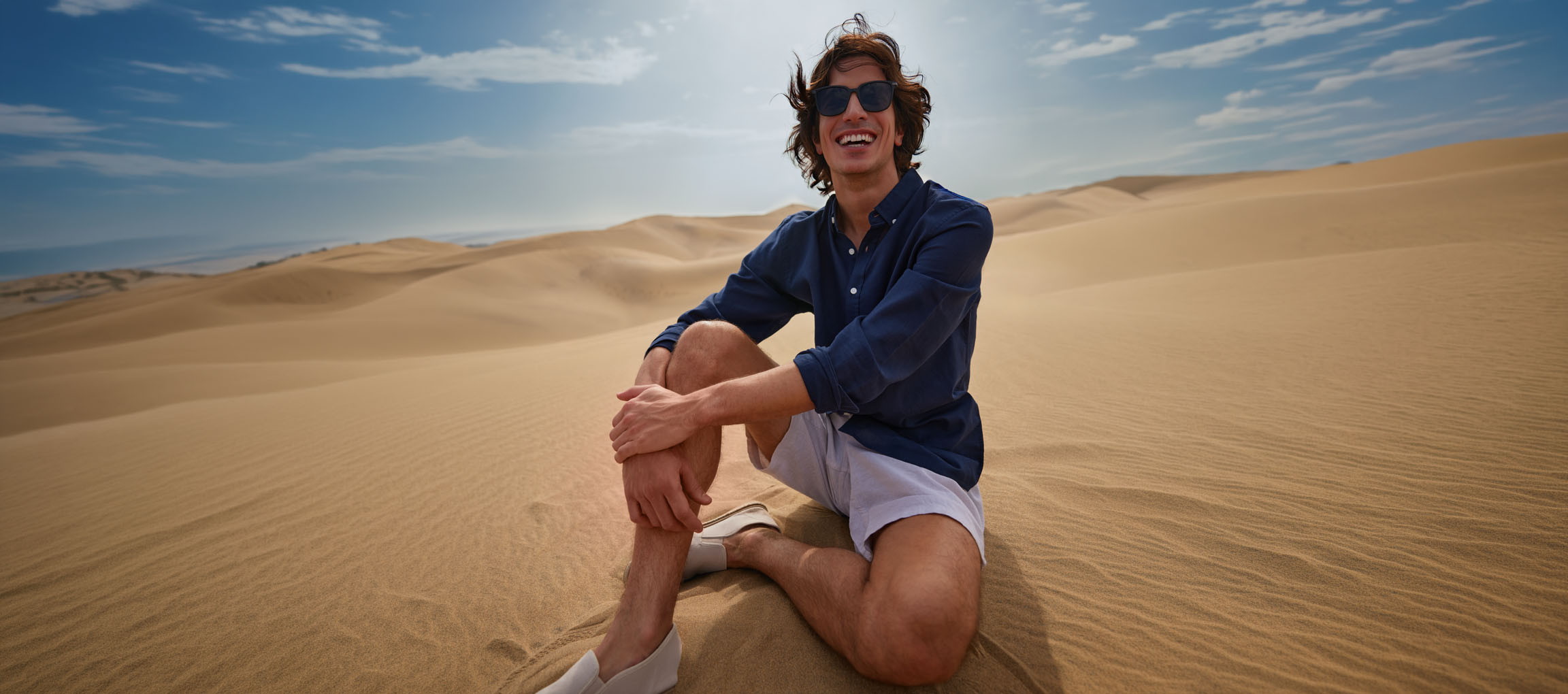 Mand i mørkeblå skjorte og shorts sidder i sandklitter og smiler.