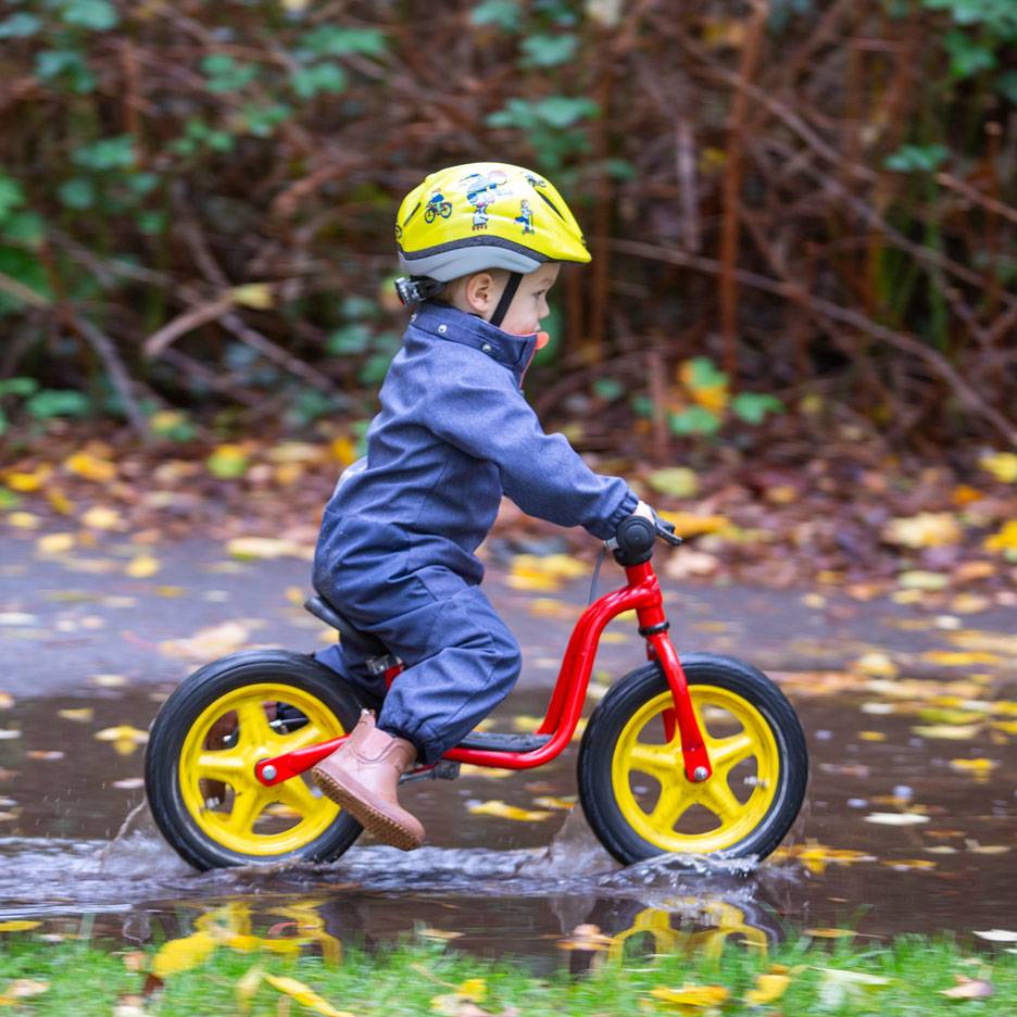 Barn på løbecykel i vandpyt, med hjelm og heldragt.