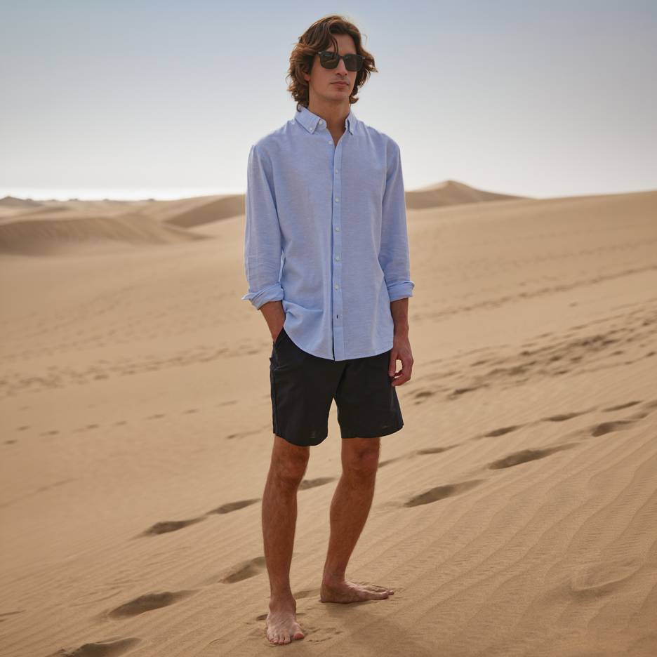 Mand i blå skjorte og sorte shorts, barfodet stående på sandklitter.