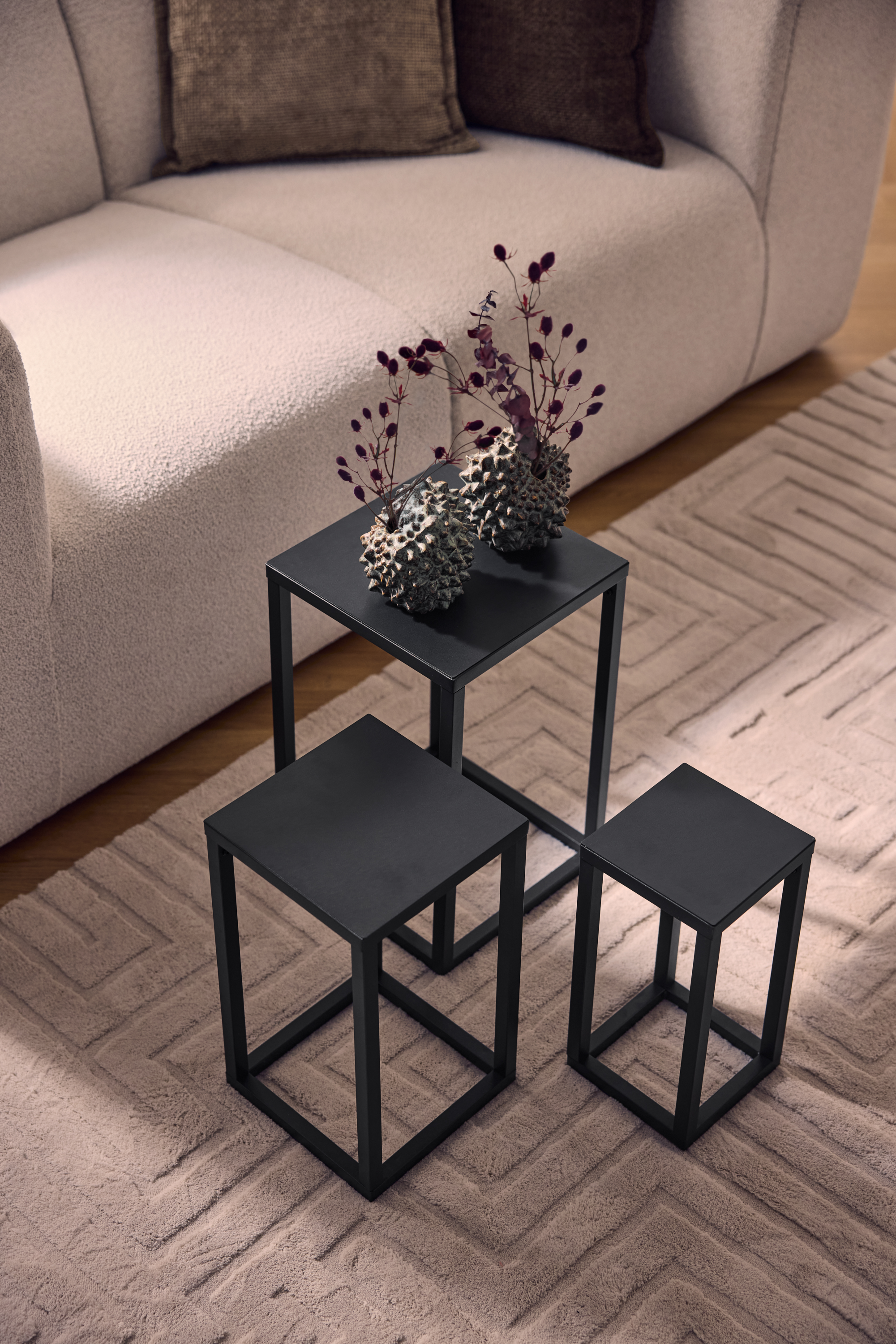 Tre sorte metal sofaborde på et beige tæppe med geometrisk mønster, ved siden af en lys sofa.