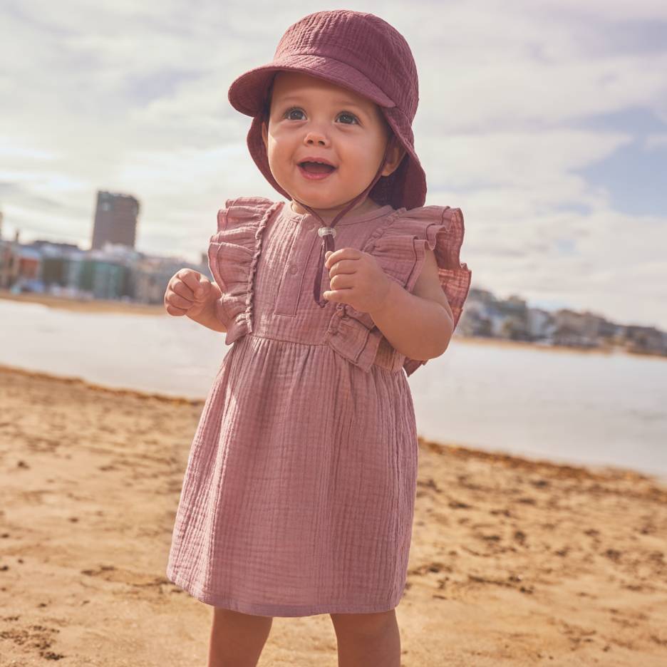 Baby i lyserød kjole og hat på stranden.