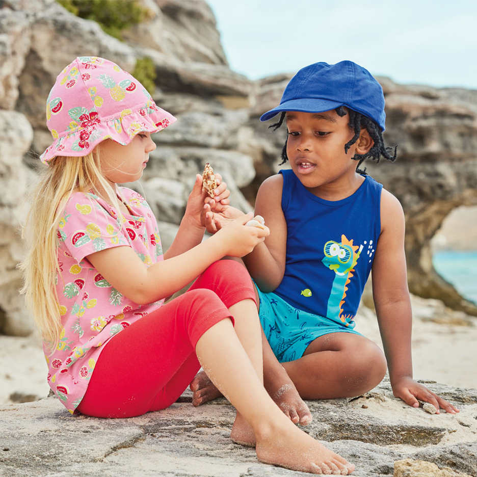 To børn på en strand, det ene i en lyserød solhat og frugtmønstret top, det andet i en blå kasket og dinosaur-tanktop, holder muslingeskaller.