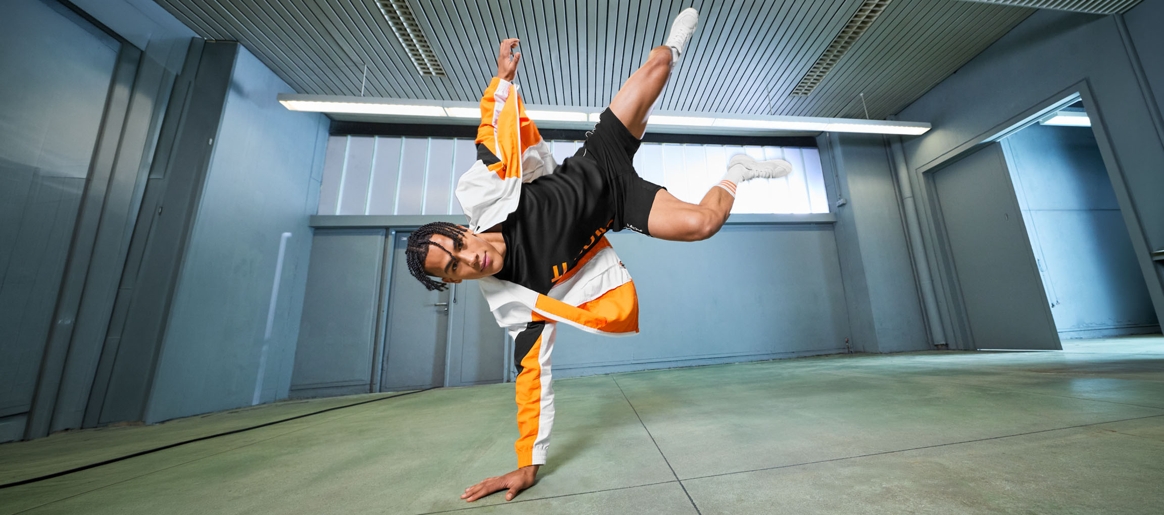 Mand i sportstøj, sort t-shirt og orange og hvid jakke, danser i et rum.