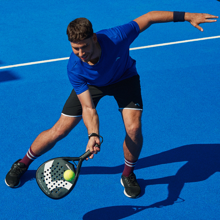 Mand i blå t-shirt og sorte shorts spiller padel med bat og bold.