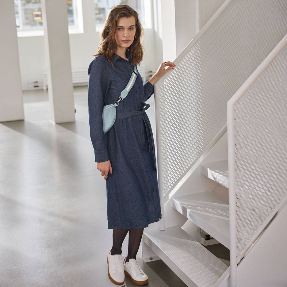 Kvinde i denim skjortekjole, sorte strømpebukser og hvide sneakers, med en lyseblå bæltetaske.