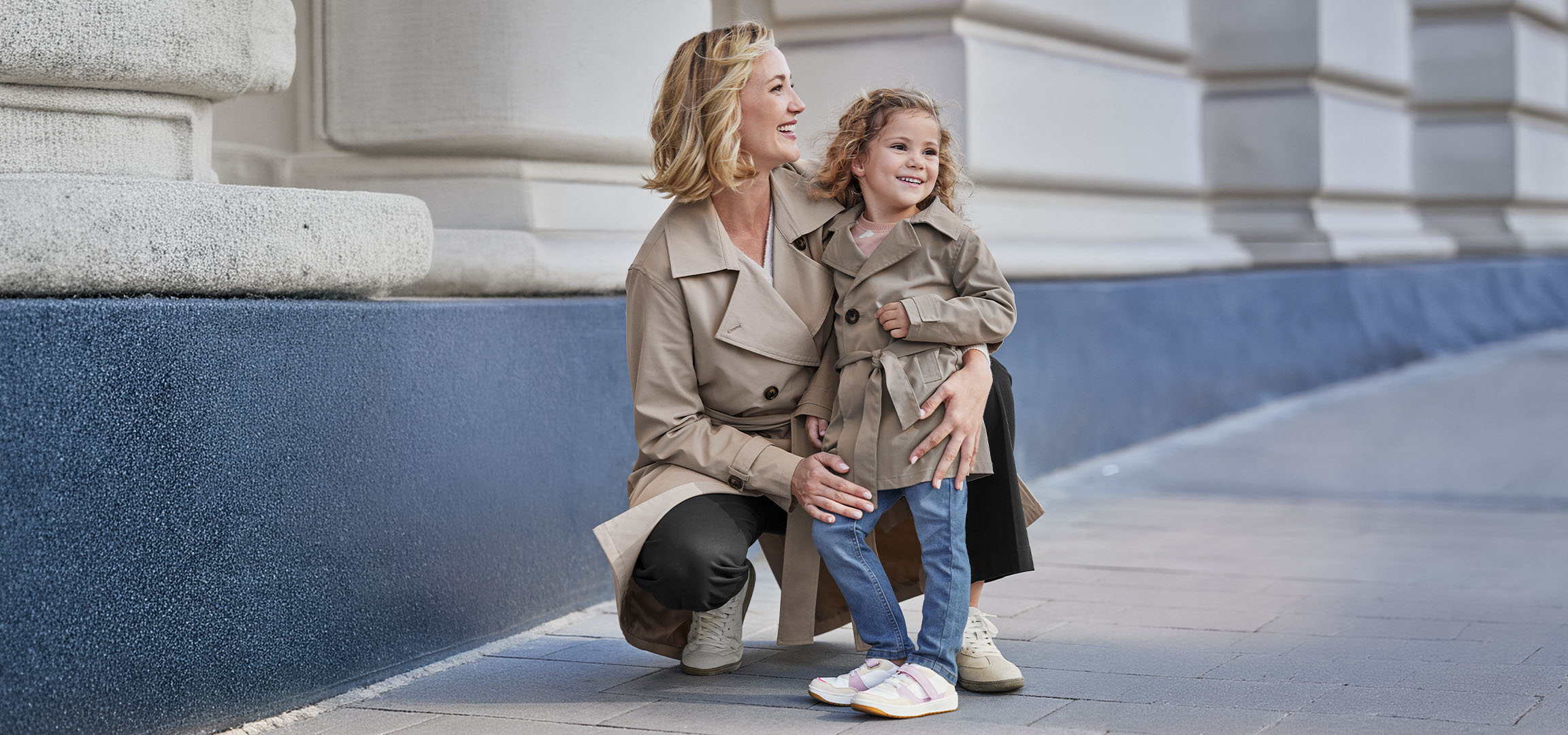 To kvinder i beige trenchcoats, en voksen og et barn, smilende udendørs.