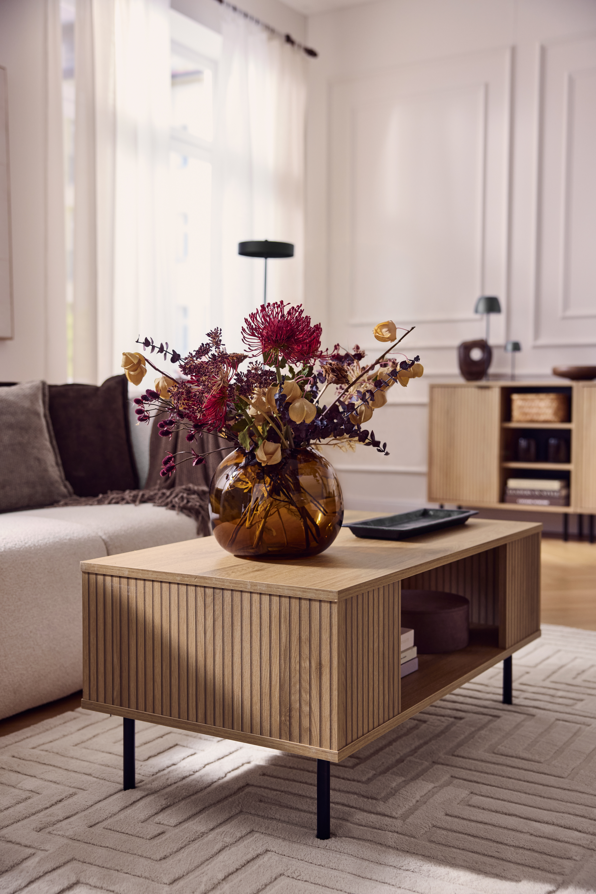 Træsofabord med riflet front, vase med tørrede blomster, beige sofa.